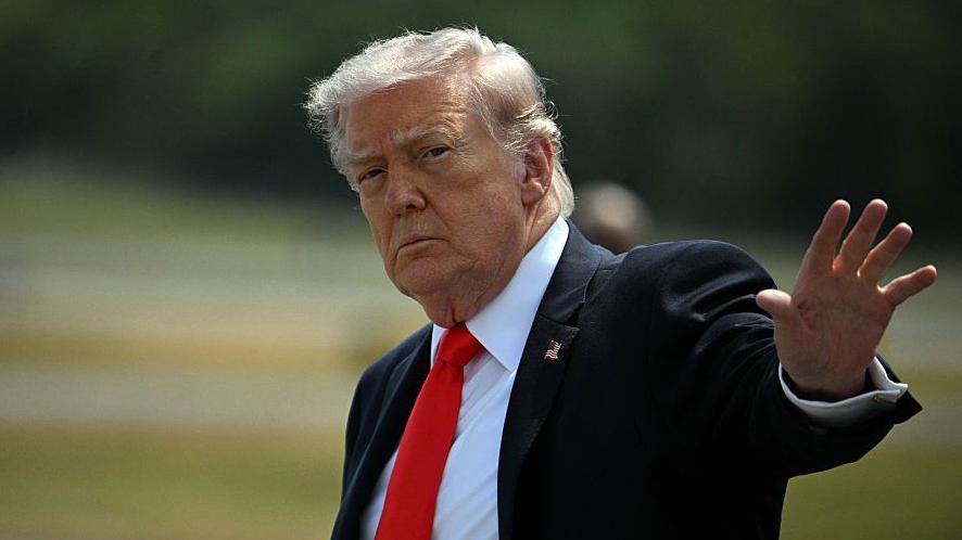 Дональд Трамп, одетый в костюм, поднимает руку в сторону камеры.