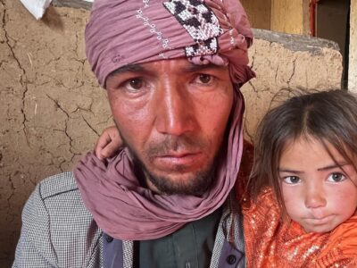 Мужчина в розовом тюрбане крепко прижимает к себе свою маленькую дочь перед потрескавшейся глинобитной стеной