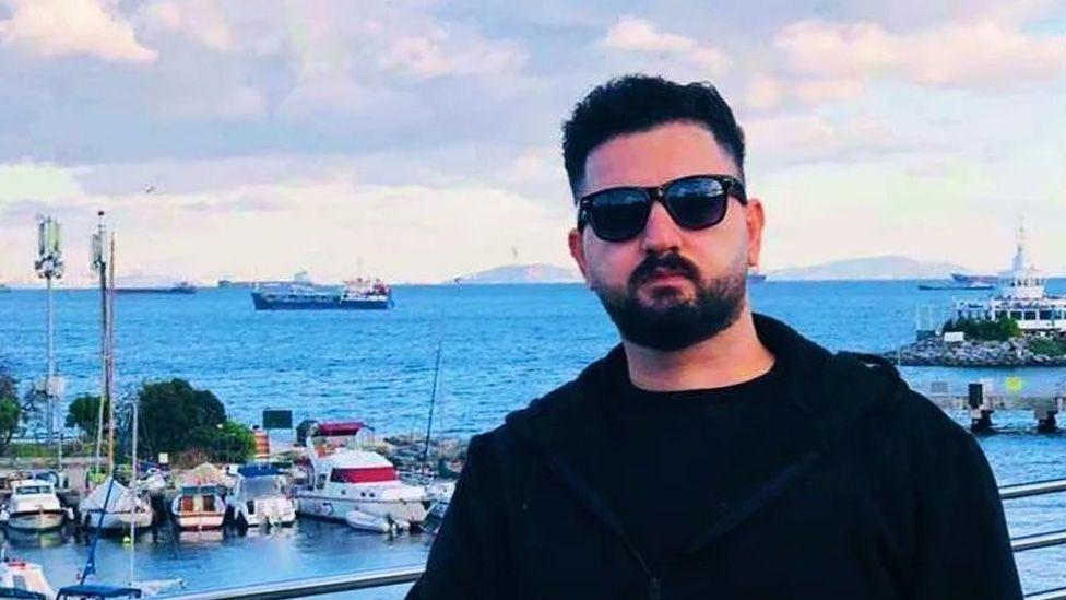 Kardo Ranya stands in the foreground on a waterfront balcony, wearing dark sunglasses, a black jacket, and a dark shirt. Behind him is a coastal marina with several small boats and yachts moored along docks, their white hulls contrasting with the deep blue water. Farther out on the sea, a few large cargo ships are visible near the horizon. To the right, a pier or harbour structure extends into the water, and low buildings line parts of the shore.