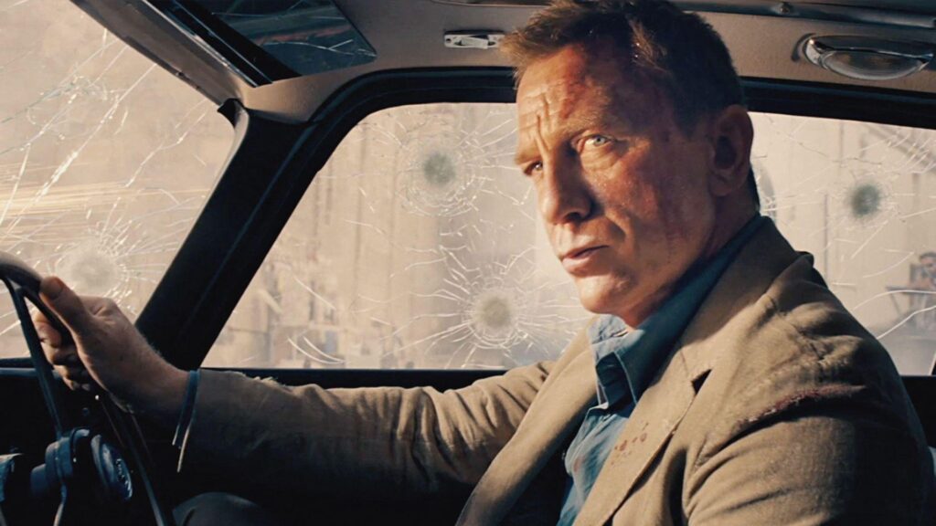 Daniel Craig sits in a car covered in gunshot impacts as he plays James Bond in the 2020 film No Time to Die