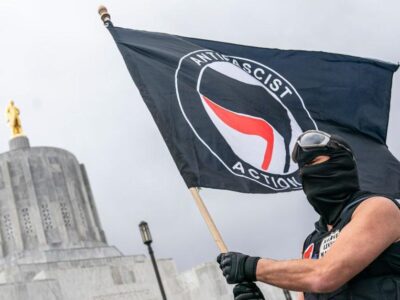 Суд в Техасе приговорил восемь активистов «Антифа» к крупным срокам за нападение на иммиграционный центр. Лидер группы получил 100 лет тюрьмы 5 Мужчина в черной балаклаве держит в руках флаг антифа