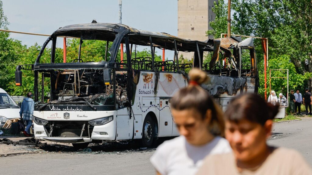В самопровозглашенной ДНР заявили о восьми погибших при ударе по рейсовому автобусу. Что известно 1 Две женщины идут по улице, за ними — сгоревший автобус.