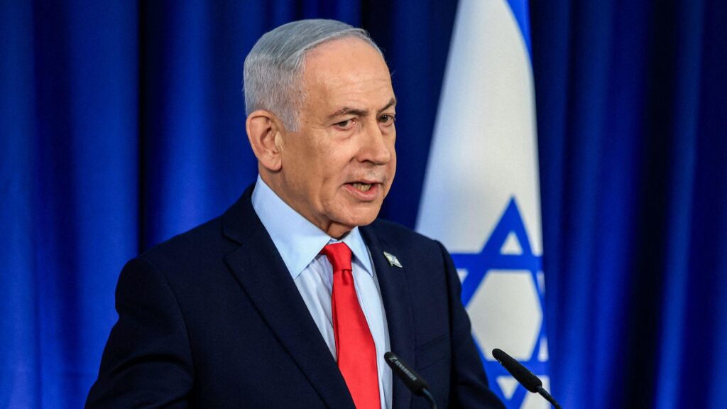 Netanyahu speaking in front of a a blue background and Israeli flag wearing a black suit and red tie