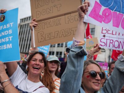 В Белфасте тысячи людей вышли на митинг против расизма после беспорядков из-за нападения с ножом 2 Женщины держат плакаты с надписями «Белфаст против расизма», «Одна любовь» и «Беспорядки не отражают позицию Белфаста». Они находятся в городском районе, над ними сгущаются серые тучи.