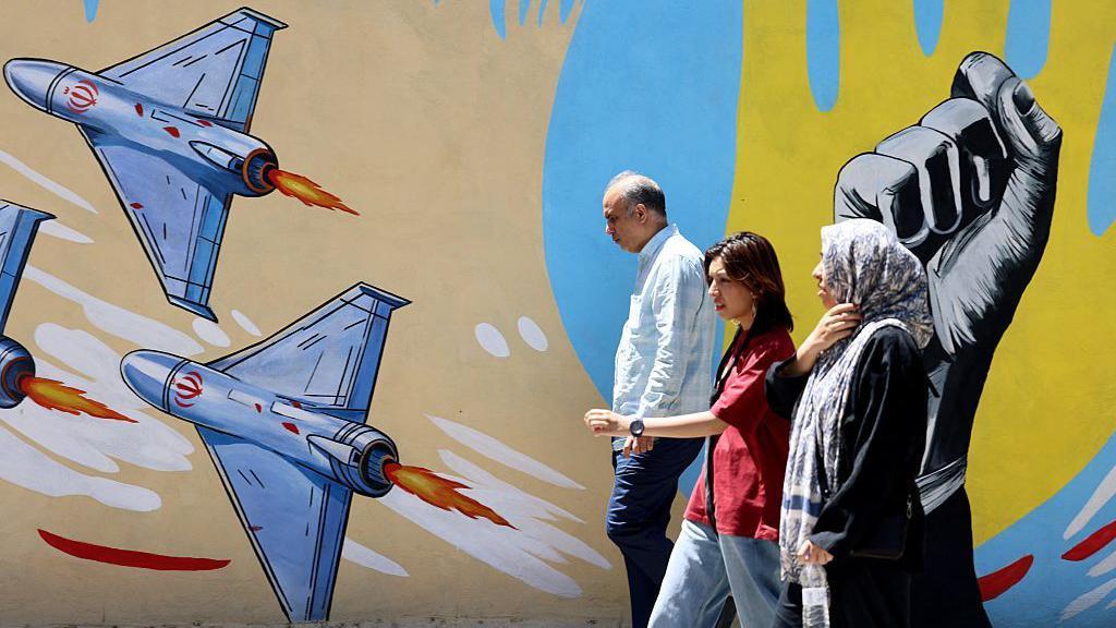 Джереми Боуэн: Трампу нужно, чтобы эта война закончилась, но Иран не сдается 1 Пожилой мужчина и женщина, а также молодая девушка проходят мимо граффити на котором изображены несколько дронов и поднятый кулак