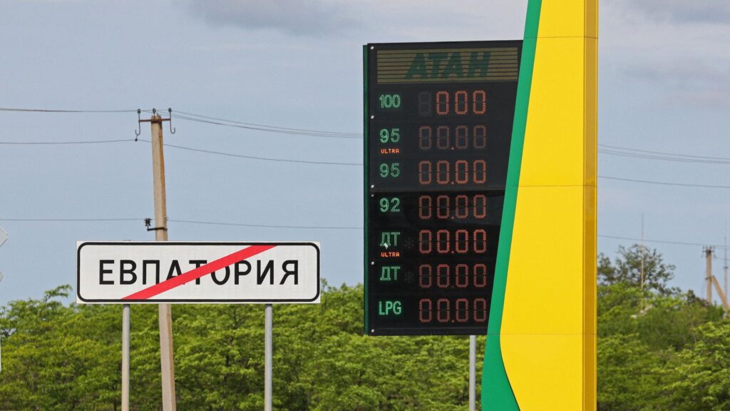 Локальный дефицит бензина. Как атаки на нефтяную инфраструктуру России повлияли на рынок топлива 1 На заднем фоне темно-синее небо, зеленые деревья и столбы с проводами. На переднем плане слева дорожный знак о выезде из Евпатории и погасшее табло автозаправки "Атан".