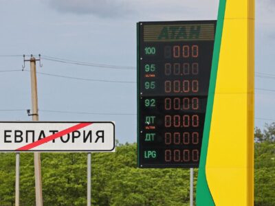 Локальный дефицит бензина. Как атаки на нефтяную инфраструктуру России повлияли на рынок топлива 2 На заднем фоне темно-синее небо, зеленые деревья и столбы с проводами. На переднем плане слева дорожный знак о выезде из Евпатории и погасшее табло автозаправки "Атан".