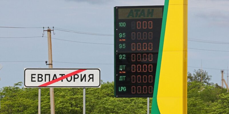На заднем фоне темно-синее небо, зеленые деревья и столбы с проводами. На переднем плане слева дорожный знак о выезде из Евпатории и погасшее табло автозаправки "Атан".