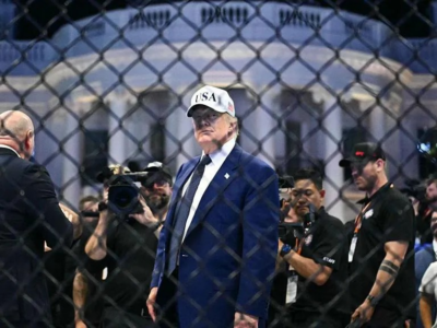За забором перед Белым домом стоит Трамп в пиджаке и белой кепке с надписью USA, за ним люди с камерами и другие люди.