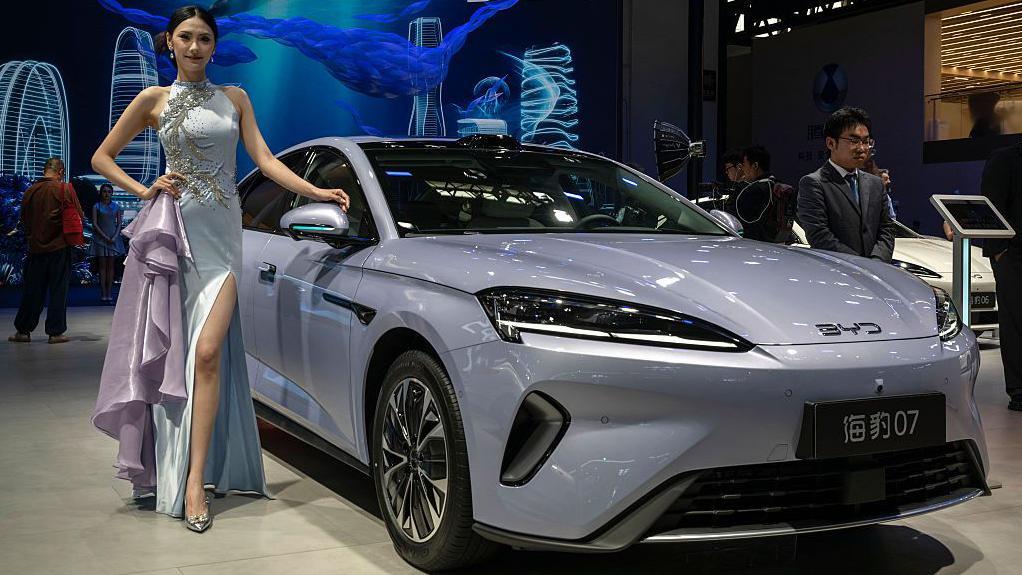 Десятки китайских компаний, включая BYD и Alibaba, попали в список Пентагона как «связанные с военными КНР» 1 Женщина в серебристом вечернем платье стоит рядом с серым электромобилем китайского автопроизводителя BYD, представленным на пресс-конференции в рамках Пекинской международной автомобильной выставки.