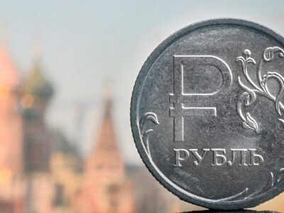 На заднем плане размытые очертания православного собора Василия Блаженного в Москве. На переднем плане монета из серого металла с надписью "рубль"