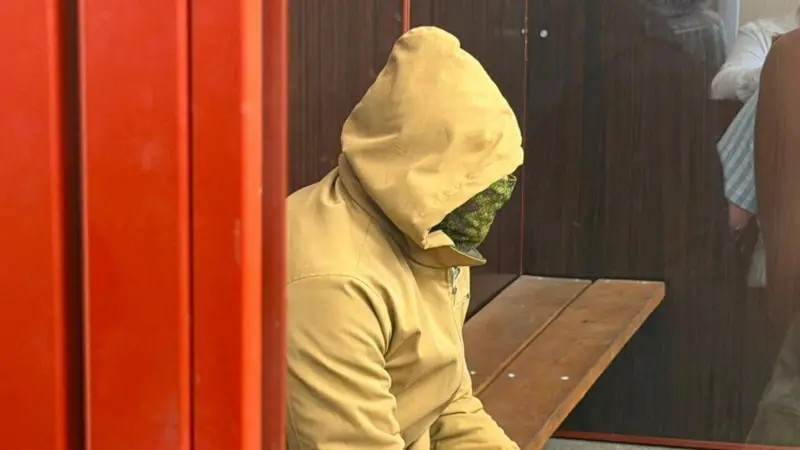 Покушение на бизнесмена Ермолаева в Монако: суд в Киеве арестовал сотрудника ГУР, подозреваемого в убийстве Березовской 1 Человек в светлом худи с капюшоном, лицо закрыто зеленой тканью. Он сидит на скамейке в стеклянном боксе в суде.