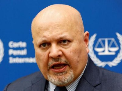 Прокурор Международного уголовного суда Карим Хан выступает во время интервью, сидя перед синей табличкой с размытыми белыми надписями и символом МУС — весами правосудия, окруженными лавровым венком.