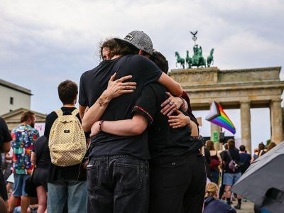 Три человека обнимают друг друга; на заднем плане виден радужный флаг ЛГБТ, а позади — Бранденбургские ворота