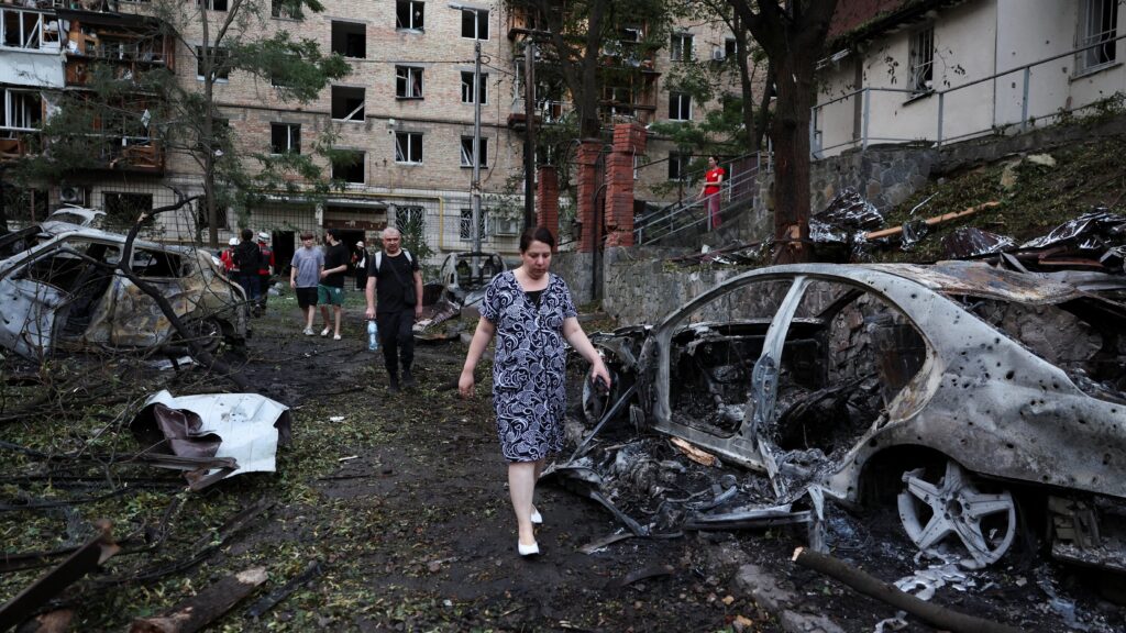 Россия нанесла массированный удар по Украине ракетами и дронами: в Киеве более 10 погибших 1 Люди идут между сгоревших машин, за их спинами поврежденный жилой дом
