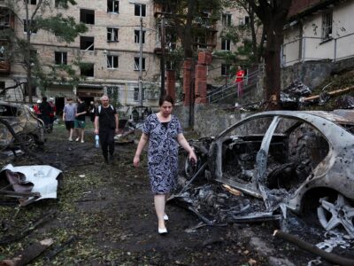 Люди идут между сгоревших машин, за их спинами поврежденный жилой дом
