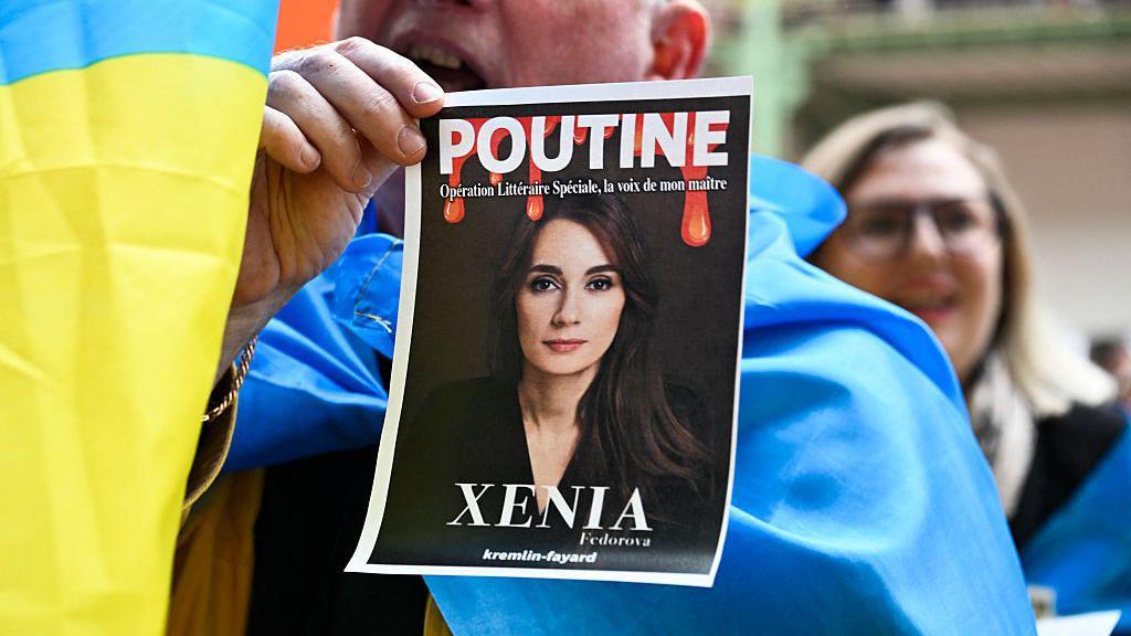 Сторонники Украины устроили акцию протеста на презентации книги Ксении Федоровой в прошлом апреле в Париже - участник акции держит фото Федоровой с надписями "Путин" и "Ксения"