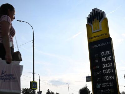 «Это и есть вторая волна». Как в России развивается топливный кризис 2 На заднем фоне — голубое небо. На среднем плане деревья, фонарные столбы и заправка «Роснефти». На табло АЗС нули напротив всех видов топлива, кроме дизельного. На переднем плане слева направо идет женщина с пакетом руке.