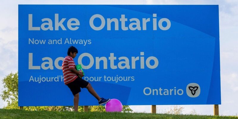 A sign reads "Lake Ontario now and always" with a boy playing with a ball nearby.