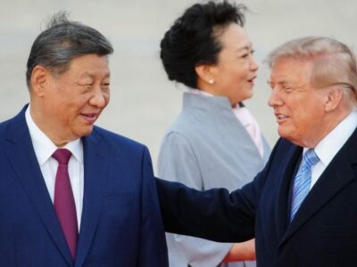President Xi smiles and looks toward Trump, who has his hand on Xi's shoulder. Both are in suits and their haor is slightly tussled by wind. Xi's wife, in a light blue coat, looks away in the background.
