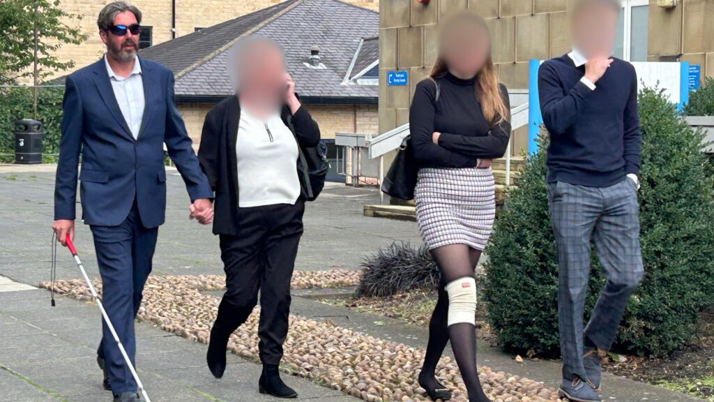В Британии слепого человека приговорили за вождение в нетрезвом виде. Он проехал за рулем 240 км 1 Четыре человека идут возле здания суда, лица троих размыты. Четвёртый человек, мужчина с бородой в синем костюме, белой рубашке и солнцезащитных очках, идёт с белой тростью