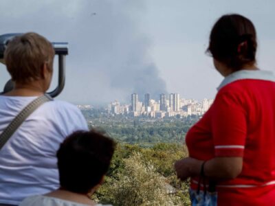 120 часов террора. Как и зачем Россия запускает волны реактивных дронов по Киеву и что Украина готовится с этим делать 4 Три женщины смотрят с возвышенности на большой столб дыма вдали, за районом многоэтажных зданий