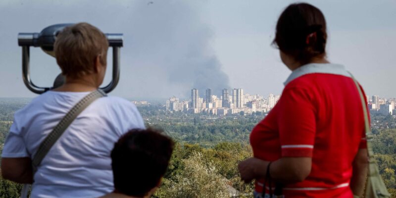 Три женщины смотрят с возвышенности на большой столб дыма вдали, за районом многоэтажных зданий