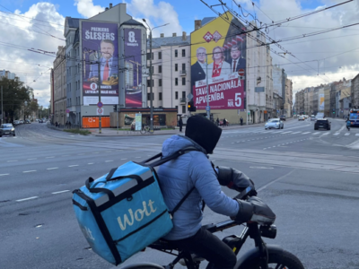 Правящие реформисты, неизбежность коалиции и «русский вопрос». В Латвии проходят парламентские выборы 2 Курьер на электровелосипеде и с контейнером за плечами на перекрестке, на заднем плане видны дома с огромными предвыборными плакатами