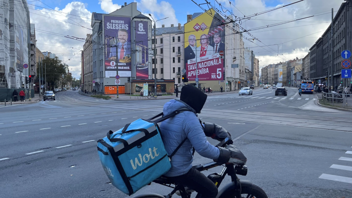 Правящие реформисты, неизбежность коалиции и «русский вопрос». В Латвии проходят парламентские выборы 1 Курьер на электровелосипеде и с контейнером за плечами на перекрестке, на заднем плане видны дома с огромными предвыборными плакатами