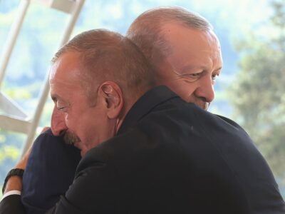Двое улыбающихся мужчин крепко обнимают друг друга - один из них Ильхам Алиев в темном костюме с усами, прижался щекой к плечу другого, президента Турции Эрдогана, тоже в темном костюме, который смотрит вниз