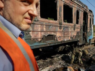 Шесть тысяч эвакуаций за месяц. Как украинские железные дороги справляются с российской кампанией ударов по ним 13 A man wearing a blue shirt and orange high vis jacket stands in front of a burnt train carriage