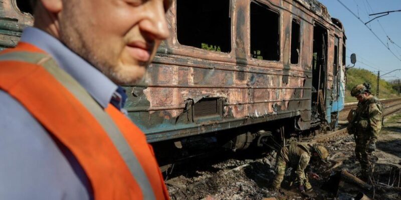 A man wearing a blue shirt and orange high vis jacket stands in front of a burnt train carriage